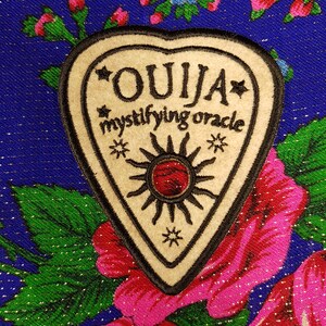 Może przedstawiać: Beżowa naszywka w kształcie planchette Ouija z czarnym obramowaniem i tekstem, w tym "OUJA" i "mystifying oracle". Planchette ma wzór słońca z czerwonym środkiem. Naszywka jest umieszczona na niebieskim tle kwiatowym.