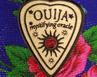 May include: A beige Ouija planchette patch with black trim and text, including "OUIJA" and "mystifying oracle." The planchette features a sun design with a red center. The patch is set against a blue floral background.