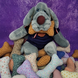 Puede incluir: Un peluche de perro gris con una nariz marrón y una pajarita estampada, vistiendo una camisa azul marino. El perro está rodeado de varios juguetes con forma de hueso de tela en diferentes colores y patrones, todo sobre un fondo morado.