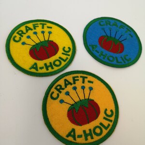 May include: Three circular patches with the text "CRAFT-A-HOLIC". The patches feature a red tomato pin cushion with blue pins. Two patches are yellow with green trim, and one is blue with green trim.