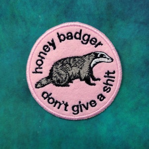 May include: A round, pink embroidered patch with a honey badger illustration. The text "honey badger don't give a shit" is embroidered around the badger. The badger is gray, black, and white.