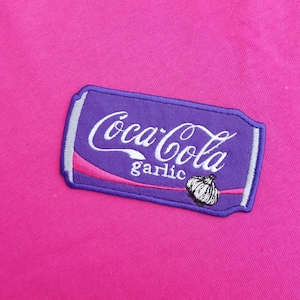 May include: A purple patch with the Coca-Cola logo in white script, the word "garlic" below, and a garlic bulb illustration. The patch is on a bright pink fabric.