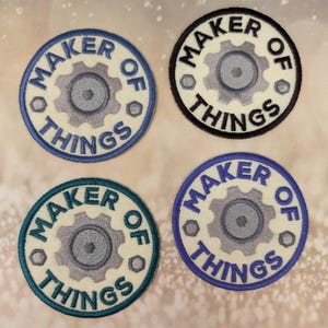 Op de afbeelding: Vier ronde geborduurde patches met de tekst "MAKER OF THINGS" in een cirkel rond een tandwiel. De patches hebben verschillende kleurenschema's: blauw, zwart, blauwgroen en paars. De achtergrond is gebroken wit.