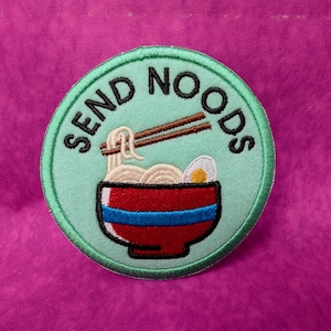 May include: A circular embroidered patch with a light mint green background featuring a red bowl of ramen noodles with a blue stripe, a soft-boiled egg, and brown chopsticks. The text SEND NOODS is embroidered in black along the top edge.