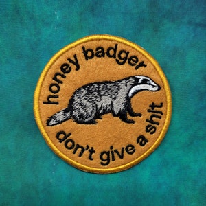 Pode incluir: Patch bordado com um desenho de texugo-do-mel em preto, branco e cinza, sobre um fundo bege. O texto "honey badger don't give a shit" está arqueado ao redor do texugo. O patch tem uma borda dourada e está em um fundo azul-petróleo.