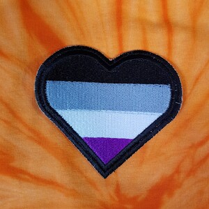 May include: A heart-shaped patch with the asexual pride flag design. The patch features a black border and a color scheme of black, gray, white, and purple. The patch is set against an orange tie-dye fabric background.