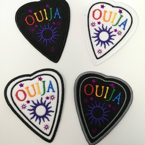 May include: Four heart-shaped Ouija patches. Two are black with rainbow text and a sun design, and two are white with the same design. The word "OUJIA" is embroidered in rainbow colours. The sun and star designs are purple.