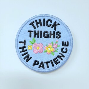 May include: A round, light blue embroidered patch with the text "THICK THIGHS THIN PATIENCE" in black. The patch features embroidered pink and yellow flowers with green leaves. The patch has a light blue border.