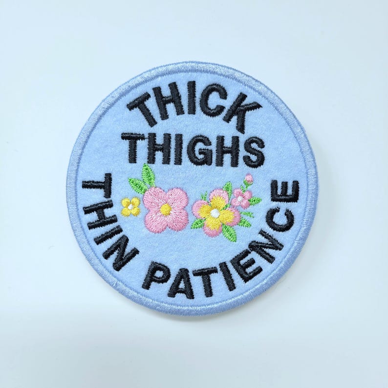 May include: A round, light blue embroidered patch with the text "THICK THIGHS THIN PATIENCE" in black. The patch features embroidered pink and yellow flowers with green leaves. The patch has a light blue border.