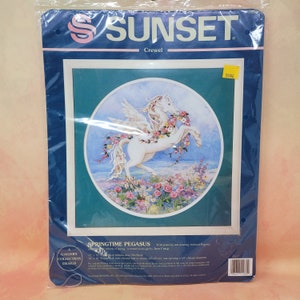 May include: A Sunset Crewel embroidery kit. The design features a white Pegasus adorned with flowers, set against a blue and green backdrop. The kit includes a framed design with the text "SPRINGTIME PEGASUS". The design measures 38cm x 38cm.