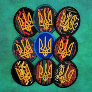 May include: A collection of embroidered patches featuring the Ukrainian Tryzub coat of arms. The patches are primarily oval-shaped with a gold emblem on various patterned backgrounds, including red, black, and blue. The patches are arranged on a green surface.