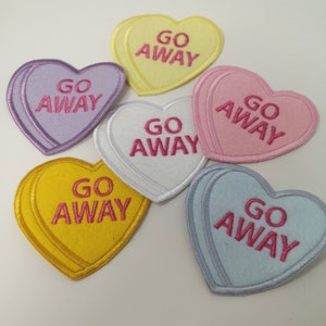 May include: A selection of heart-shaped patches in pastel shades: yellow, lilac, pink, white, and pale blue. Each patch features the embroidered phrase "GO AWAY" in pink, with a contrasting border.