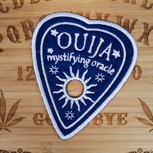 May include: A navy blue Ouija patch with white embroidered details, including the word "OUJIA" and "mystifying oracle". The patch is heart-shaped with a sun design and star accents. The patch is on a wooden surface.