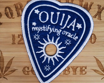May include: A navy blue Ouija patch with white embroidered details, including the word "OUJIA" and "mystifying oracle". The patch is heart-shaped with a sun design and star accents. The patch is on a wooden surface.
