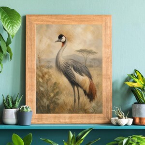 May include: Framed print of a Grey Crowned Crane in a savanna setting. The bird features grey, white, and brown plumage, a black and white face, and a golden crown. The artwork is presented in a light brown wooden frame.