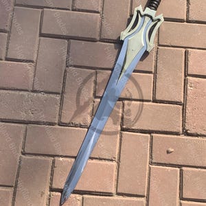 May include: A long, silver sword with a decorative hilt. The hilt is silver and black, with a stylized design. The blade is long and has a slight curve. The sword is lying on a brick surface.