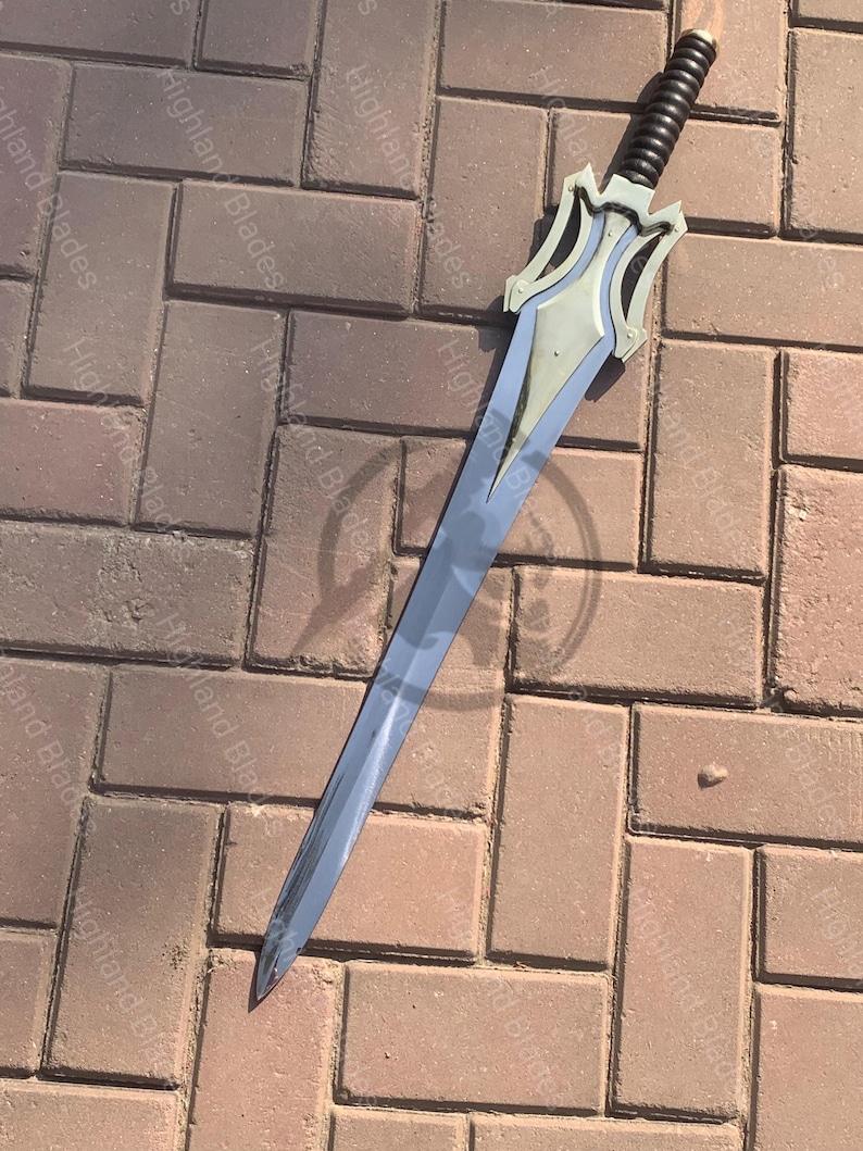 May include: A long, silver sword with a decorative hilt. The hilt is silver and black, with a stylized design. The blade is long and has a slight curve. The sword is lying on a brick surface.