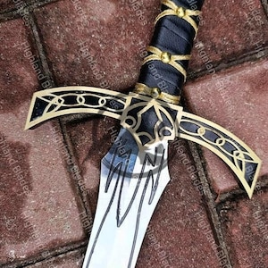 May include: Close-up of a decorative sword with a black leather-wrapped handle and gold accents. The blade is silver with etched designs. The crossguard and pommel feature intricate gold detailing. The sword rests on a brick surface.