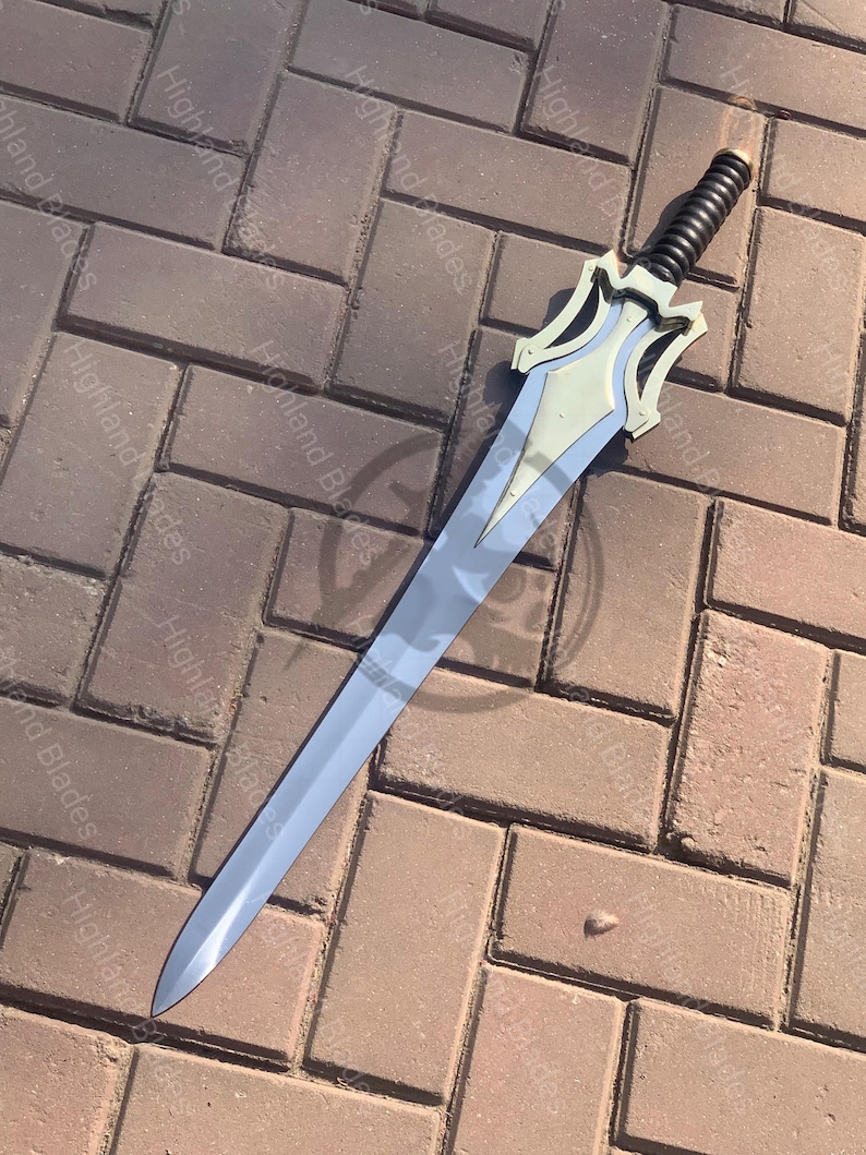 May include: A silver-colored sword with a decorative hilt and a black handle. The blade is long and slender, with a pointed tip. The hilt has a stylized design with a light gold color. The sword is lying on a brick surface.