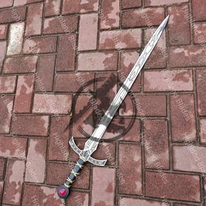 May include: A silver sword with a decorative handle featuring a black grip, gold accents, and a red gem. The blade has etched designs. The sword is lying on a brick surface.