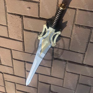 May include: A decorative dagger with a silver blade and a gold-colored hilt. The handle is black with a ridged design. The dagger is lying on a brick surface. The text "Highland Blades" is visible on the surface.