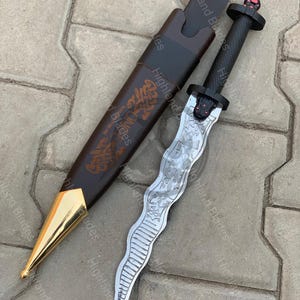 Puede incluir: Una daga decorativa con una hoja plateada ondulada y un mango negro. La daga se muestra junto a su funda de cuero marrón, que tiene una punta dorada y un diseño en relieve. El texto "Highland Blades" es visible.