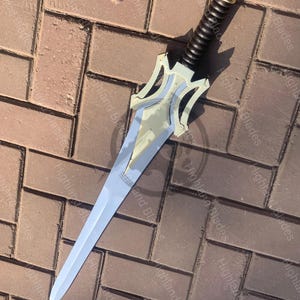 May include: A silver and gold sword with a black handle. The blade is long and pointed, with a decorative hilt featuring a stylized design. The handle is wrapped in a textured material. The sword is lying on a brick surface.