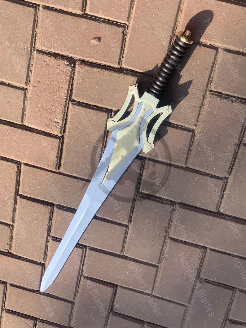 May include: A silver and gold sword with a black handle. The blade is long and pointed, with a decorative hilt featuring a stylized design. The handle is wrapped in a textured material. The sword is lying on a brick surface.