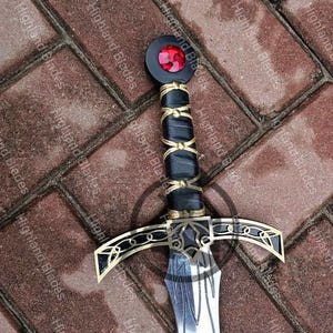 May include: A decorative sword with a black handle wrapped in gold-colored wire and leather. The hilt features gold accents and a red gem. The blade is silver with etched details. The words "Highland Blades" are printed on the background.