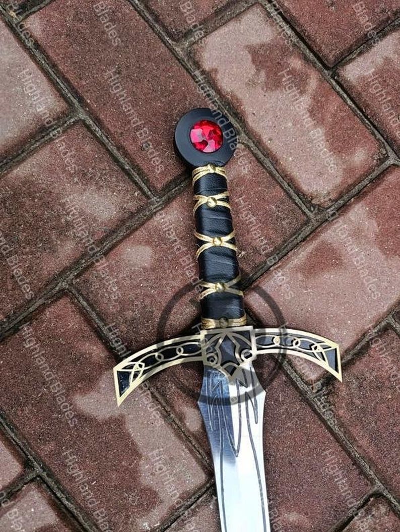 May include: A decorative sword with a black handle wrapped in gold-colored wire and leather. The hilt features gold accents and a red gem. The blade is silver with etched details. The words "Highland Blades" are printed on the background.