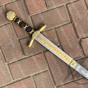 Puede incluir: Una espada decorativa con empu&ntilde;adura y guarda doradas, un mango envuelto en negro y dorado y una hoja plateada. La hoja presenta letras doradas. La espada se exhibe sobre una superficie de ladrillo.