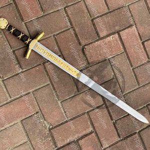 Puede incluir: Una espada decorativa con empu&ntilde;adura dorada y hoja plateada. La empu&ntilde;adura tiene un agarre envuelto en cuero negro y un pomo dorado. La hoja tiene letras doradas. La espada se exhibe sobre una superficie de ladrillo.