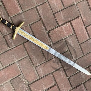 Puede incluir: Una espada decorativa con guarda y empu&ntilde;adura doradas, un mango envuelto en negro y dorado y una hoja gris claro con un dise&ntilde;o estampado. La hoja tiene texto grabado. La espada mide aproximadamente 76 cm de largo.