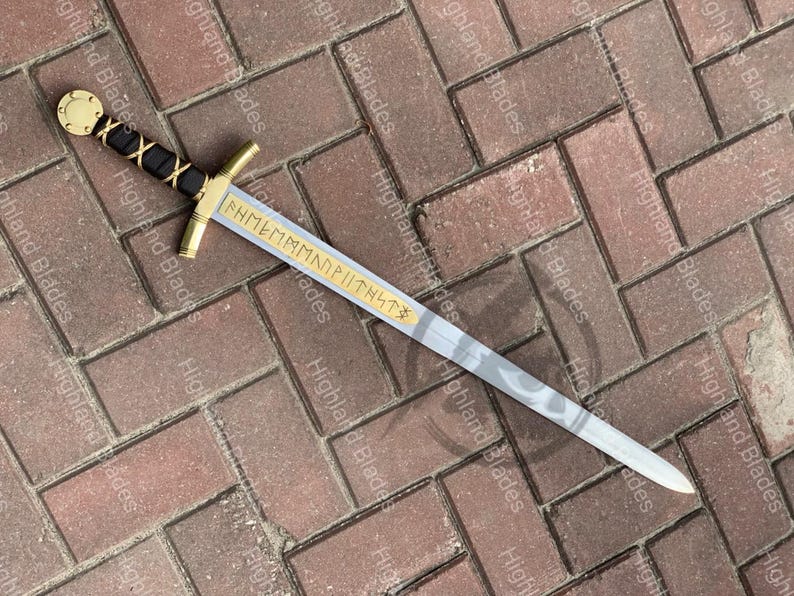 Puede incluir: Una espada decorativa con guarda y empu&ntilde;adura doradas, un mango envuelto en negro y dorado y una hoja gris claro con un dise&ntilde;o estampado. La hoja tiene texto grabado. La espada mide aproximadamente 76 cm de largo.