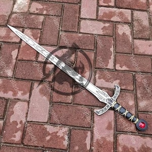 May include: A decorative sword with a silver blade and ornate handle. The handle features black and gold accents, a red gem, and a cross-shaped guard. The sword is displayed on a brick surface.