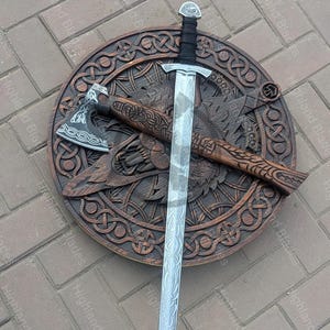 May include: A decorative wooden shield with intricate carvings, an axe, and a long sword. The sword has a silver blade with etched designs and a black handle. The axe and shield are brown. The text "Highland Blades" is visible.