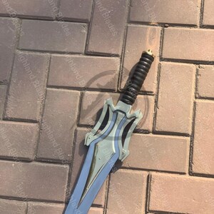 May include: A silver and blue sword with a black handle. The blade has a pointed tip and a decorative hilt. The handle is black with a ridged design. The sword is lying on a brick surface. The text "Highland Blades" is visible.