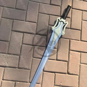 May include: A silver and blue sword with a black handle. The sword has a decorative hilt with a light blue design. The blade is long and pointed, and the sword is lying on a brick surface.