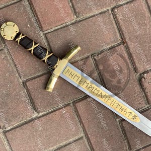 Puede incluir: Una espada decorativa con empu&ntilde;adura y guarda doradas, un mango envuelto en negro y dorado y una hoja plateada. La hoja presenta letras doradas grabadas. La espada se exhibe sobre una superficie de ladrillo.