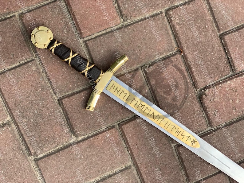 Puede incluir: Una espada decorativa con empu&ntilde;adura y guarda doradas, un mango envuelto en negro y dorado y una hoja plateada. La hoja presenta letras doradas grabadas. La espada se exhibe sobre una superficie de ladrillo.