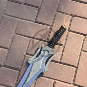 May include: A silver and gray sword with a black handle. The blade has a pointed tip and a decorative guard. The handle is wrapped in a textured black material. The sword is lying on a brick surface.