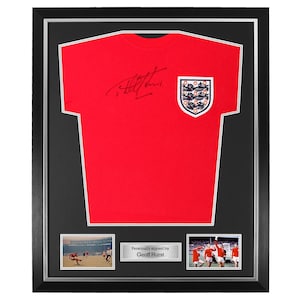 May include: Framed red England football shirt featuring the team crest and a signature. The black frame includes two small photos and the text "Personally signed by Geoff Hurst". A classic piece of memorabilia.