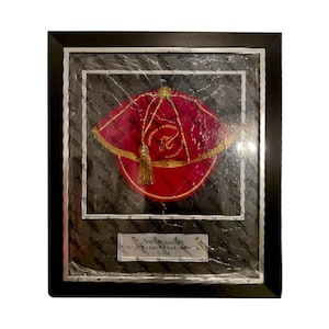 May include: Framed red and gold religious hat with a gold tassel. The hat is displayed in a black frame with a silver border. A small plaque with text is visible at the bottom.