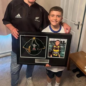 May include: A framed presentation featuring a rugby cap and a photograph. The cap is black with gold trim and a Welsh emblem. The photo shows a young person in a rugby uniform. The frame is black.