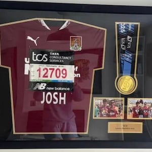 May include: Framed sports memorabilia including a maroon jersey, race bib with "JOSH" and number 12709, a medal, and two photos. The jersey has a white Puma logo and a team crest. The frame is black.