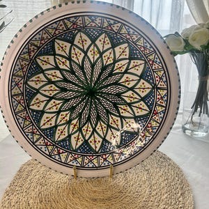 Pode incluir: Um prato de servir de cerâmica branca com um design floral detalhado em verde, azul e vermelho. O prato tem uma borda preta e é exibido em um descanso de pratos tecido. O design é simétrico e intrincado.