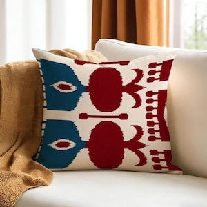 May include: Decorative throw pillow with a cream background and a repeating pattern in blue and red. The pattern features stylized shapes resembling flowers and geometric elements. The pillow is placed on a white sofa.