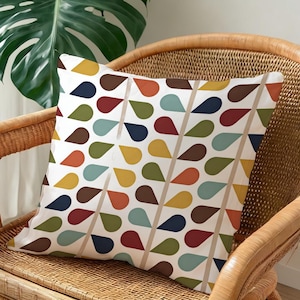 May include: Square throw pillow with a white background and a repeating pattern of colorful teardrop shapes in shades of brown, red, yellow, green, blue, and orange. The pillow is resting on a wicker chair.