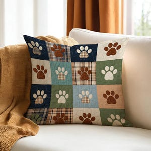 Pode incluir: Almofada decorativa quadrada com um design de patchwork com pegadas de patas. A almofada tem uma variedade de cores, incluindo azul marinho, castanho, verde e bege. As pegadas são brancas e castanhas.