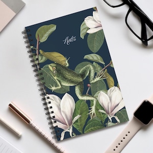 Peut inclure: Cahier à spirales avec une couverture bleu foncé ornée d'une illustration détaillée d'une grenouille verte, de fleurs de magnolia blanches et de feuilles vertes. Le mot "Notes" est écrit en cursive. Le cahier est ouvert.